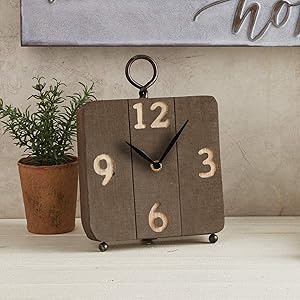 Brown wooden faced clock sits next to a potted plant on a table up against a neutral colored wall