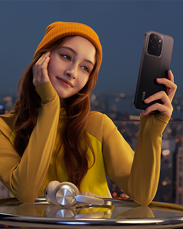 Persona con suéter amarillo y gorro naranja que se hace una selfie con un teléfono inteligente de color oscuro en un entorno interior con las luces de la ciudad visibles de fondo.