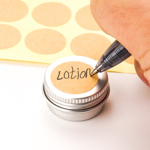 Close-up of a small metal container or jar with handwritten text on its lid, photographed against a peach-colored background.