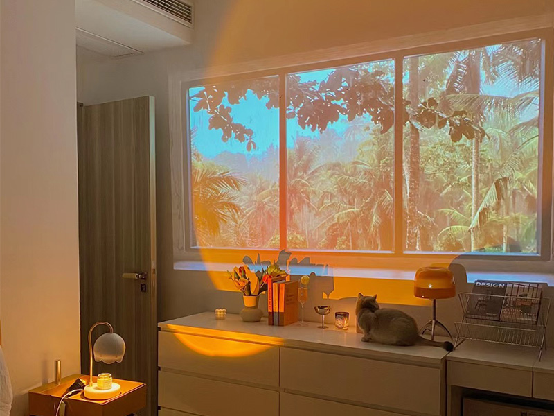 Intérieur de la pièce avec une grande fenêtre donnant sur le feuillage tropical, un éclairage d'ambiance chaleureux et des meubles, y compris une commode et une petite lampe de bureau.