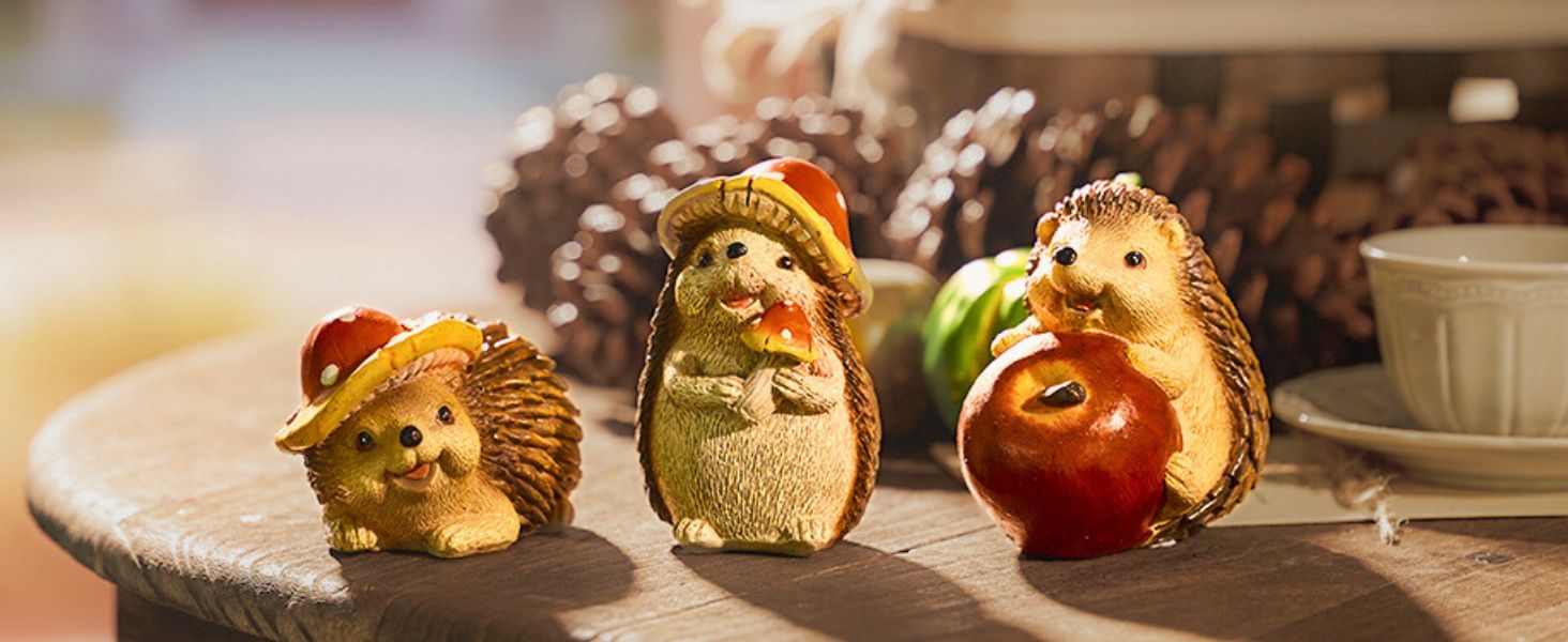 Three decorative hedgehog figurines on a wooden surface. One wears a red cap, another an orange hat, and the third holds a red apple.