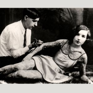Black and white photo of a man giving a woman a tattoo on her arm. She appears covered in tattoos.