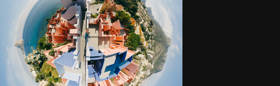 Vista aérea panorámica de 360 grados de una ciudad costera con edificios con techos rojos agrupados a lo largo de la costa, rodeados de una costa rocosa y aguas azules.