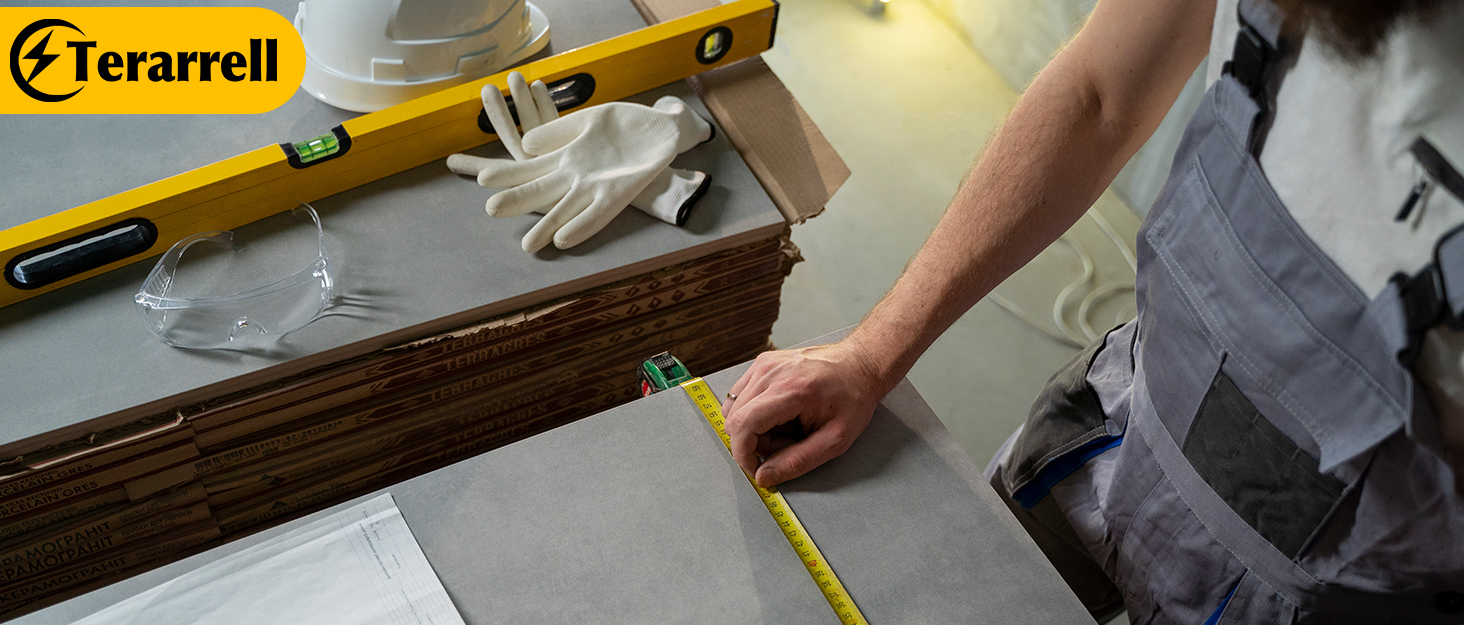 Tile installation process with hands measuring gray tiles. Yellow level, work gloves, and Terarrell brand logo visible. Demonstrates precision in tile placement.
