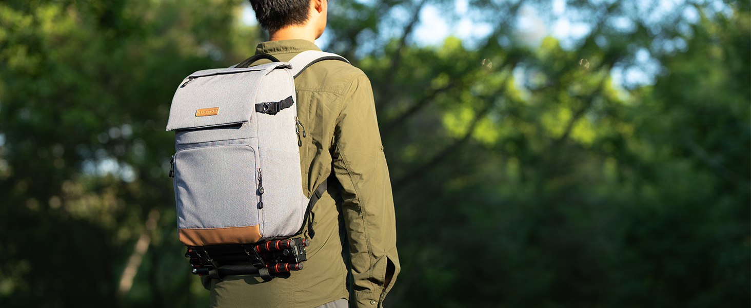 Persona che indossa uno zaino di colore chiaro con più scomparti e cinghie, in piedi in un'area boschiva con la luce del sole che filtra attraverso