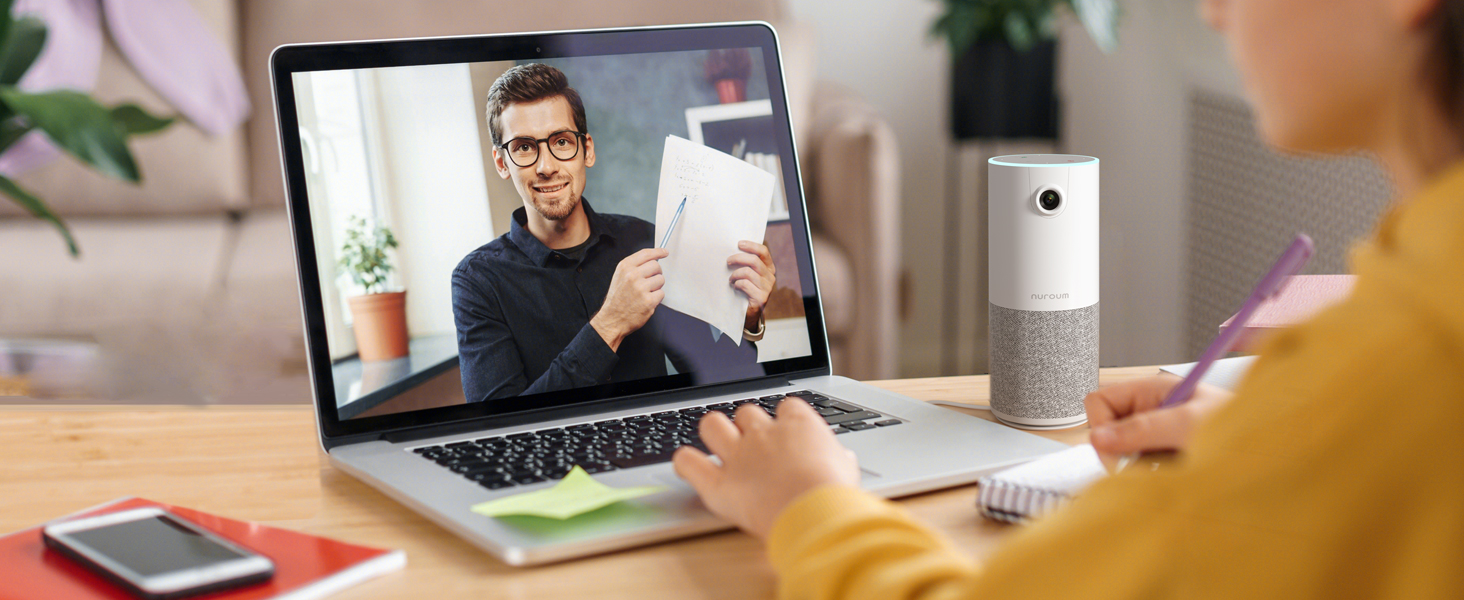 Laptop displaying video call with person holding document. Smart speaker and smartphone on desk. Person's hands typing on laptop keyboard.