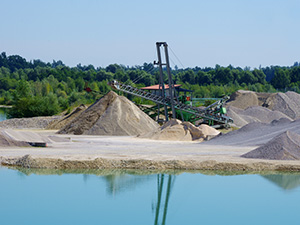 Industrieller Steinbruch oder Bergbaustandort mit Haufen von Zuschlagstoffen neben blauem Wasserkörper, im Hintergrund Förderanlagen.