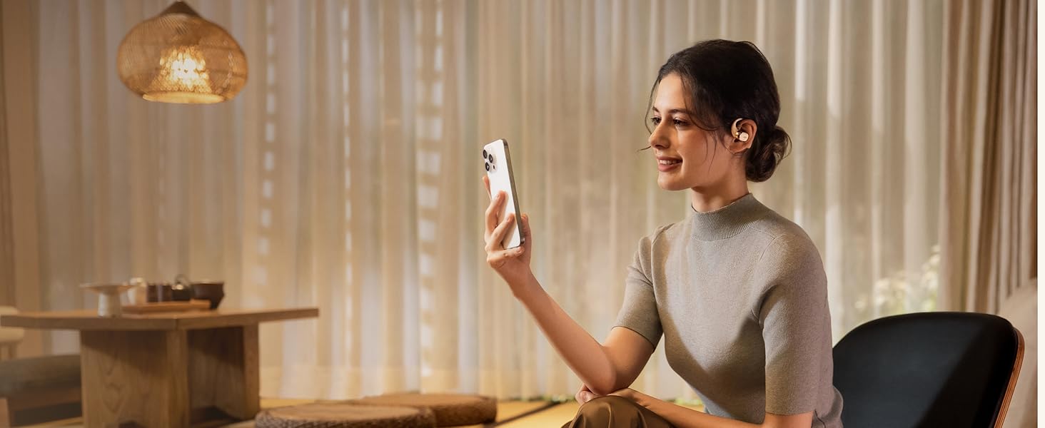 Smartphone blanc tenu par une personne en haut gris. L'appareil affiche un écran lumineux dans une pièce faiblement éclairée avec une table en bois et une lampe tissée au-dessus