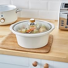 White ceramic slow cooker or crockpot with glass lid on wooden cutting board, containing what appears to be soup or stew.