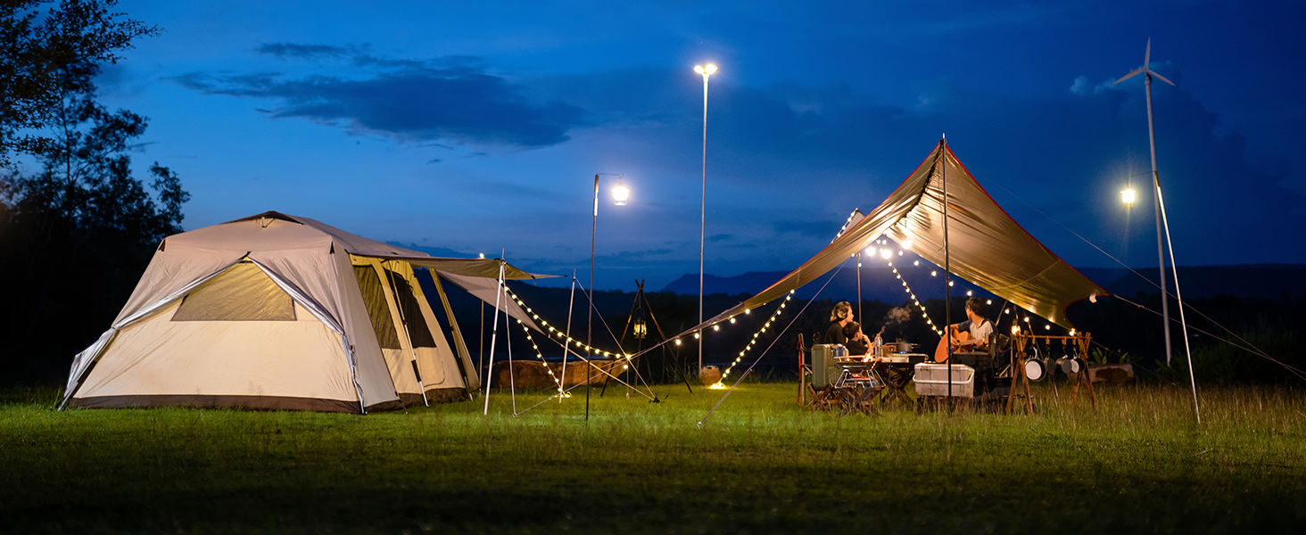 Nighttime camping scene with illuminated dome tent and large open-sided canvas tent. String lights and tall lamps provide ambient lighting. People gathered under canvas tent with tables and chairs.