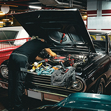 Open hood of a black classic car in a garage, revealing a complex engine compartment with various mechanical components and wiring visible.