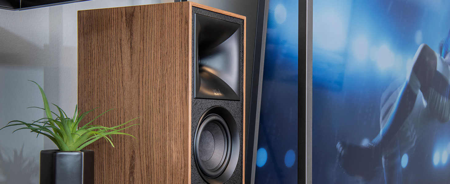 The Fives bookshelf speaker sitting on a living room bookshelf near plant and tv