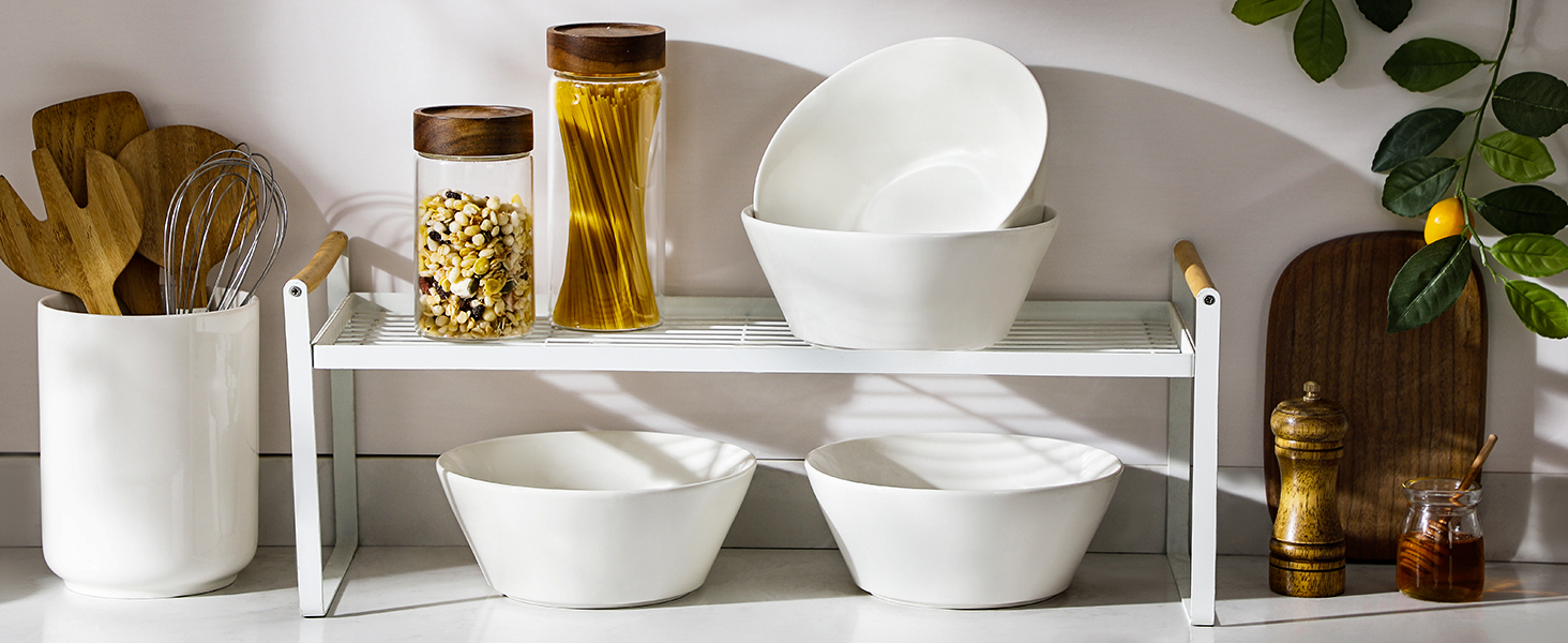 Kitchen shelf with white ceramic bowls, glass jars containing pasta, and wooden cutting boards. Minimalist modern kitchenware display on a light-colored surface.