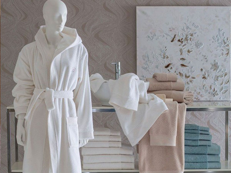 Scena da bagno con accappatoio in spugna bianca, asciugamani piegati in vari colori e una scultura o un manichino bianco parzialmente visibile