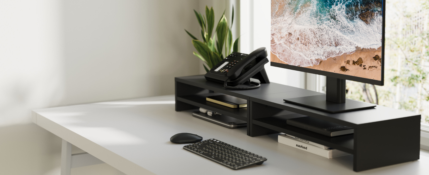 Desktop workspace setup featuring a black monitor riser shelf spanning the desk width, holding computer monitor, keyboard, and other peripherals against white wall.