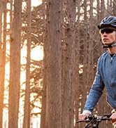 Ciclista con casco y mochila montando en bicicleta de montaña por un bosque de árboles altos iluminado por el sol