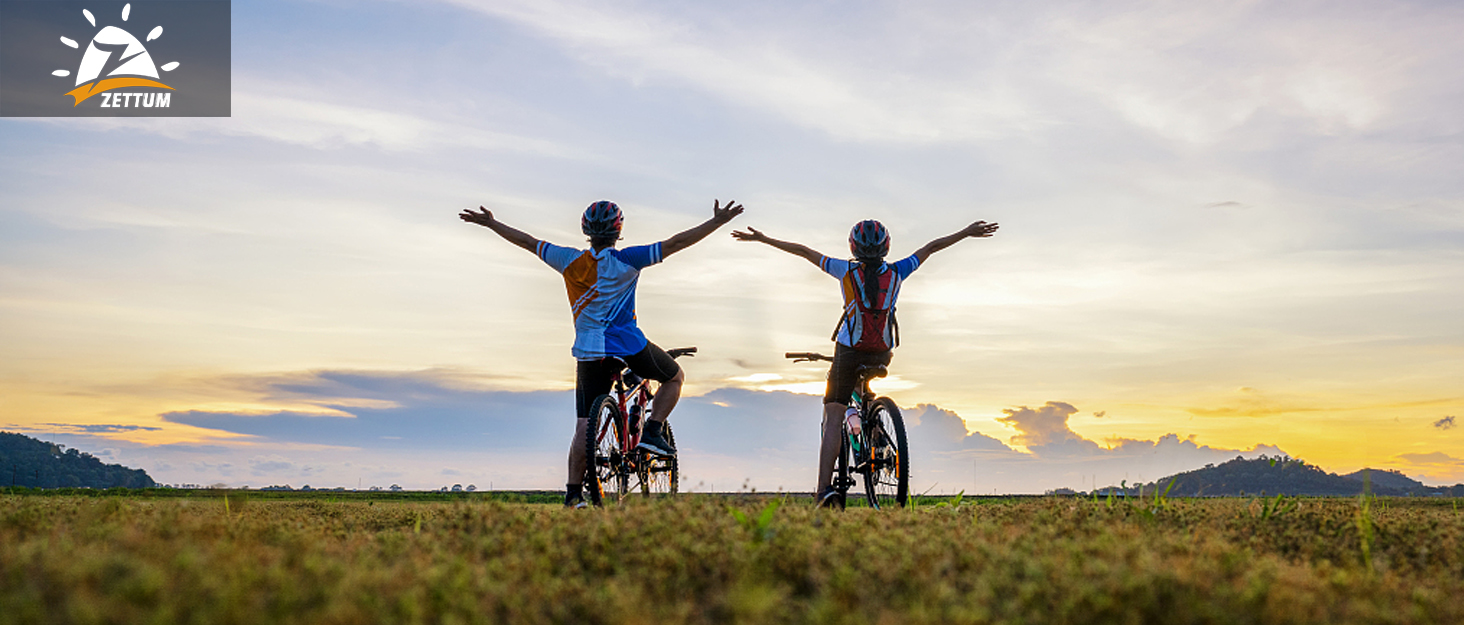 Two cyclists on mountain bikes silhouetted against a sunset sky, arms raised in celebration. Grassy field in foreground, cloudy horizon in background.