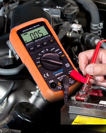 Orange digital multimeter in use, measuring voltage on a car battery. Display shows 0.05 reading.