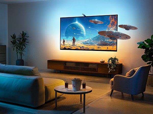 Large flat-screen TV mounted on wall displaying colorful sci-fi scene. TV emits ambient light, illuminating modern living room with couch, armchair, and wooden media console.