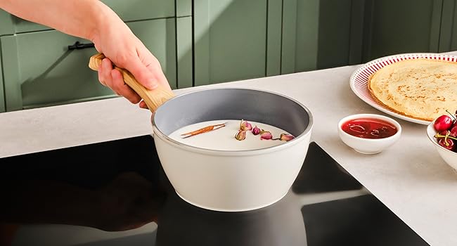 Con la mano, coge un cuenco de cerámica blanca que contiene comida caliente con vapor visible, colocado sobre una superficie de vidrio oscuro sobre una placa de cocción. Platos adicionales visibles.