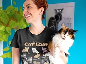 Smiling woman wearing “Types of Cat Loaf” T-shirt, holding a fluffy cat in a colorful room.