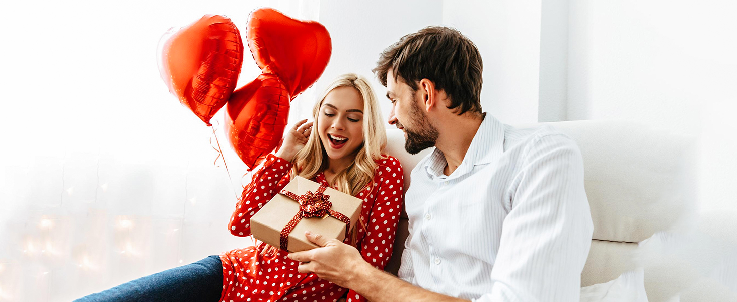 Pareja en el sofá con globos en forma de corazón. La mujer con un vestido rojo de lunares sostiene un regalo, el hombre con camisa blanca le sonríe