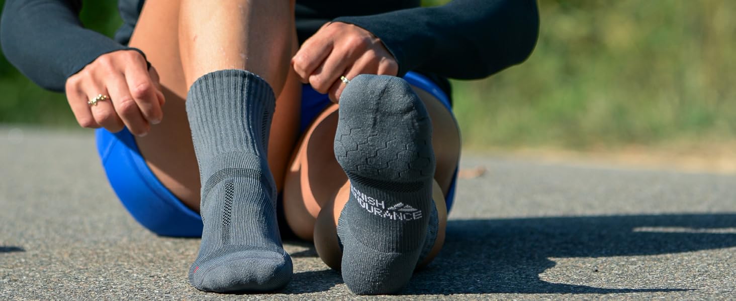 A close-up of a person sitting on a road, wearing a pair of grey DANISH ENDURANCE Cushioned Running Socks over blue shorts and a black long-sleeve top, with hands adjusting the socks. The socks feature the brand name on the soles, set against a scenic outdoor background.