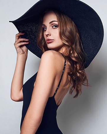 Femme portant un grand chapeau de soleil noir et un haut sans manches foncé, posant d'une main sur le bord du chapeau sur un fond