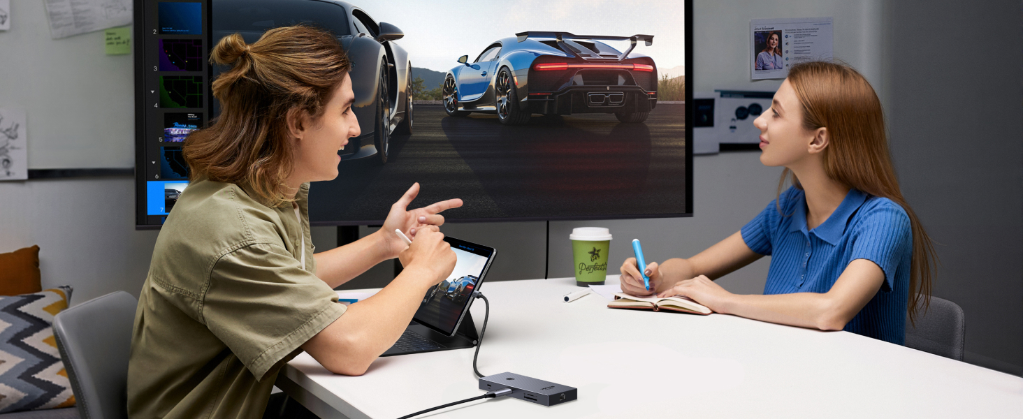 Deux personnes assises à une table dans un environnement de bureau, engagées dans une discussion. Un grand écran en arrière-plan affiche l'image d'une voiture de sport bleue.