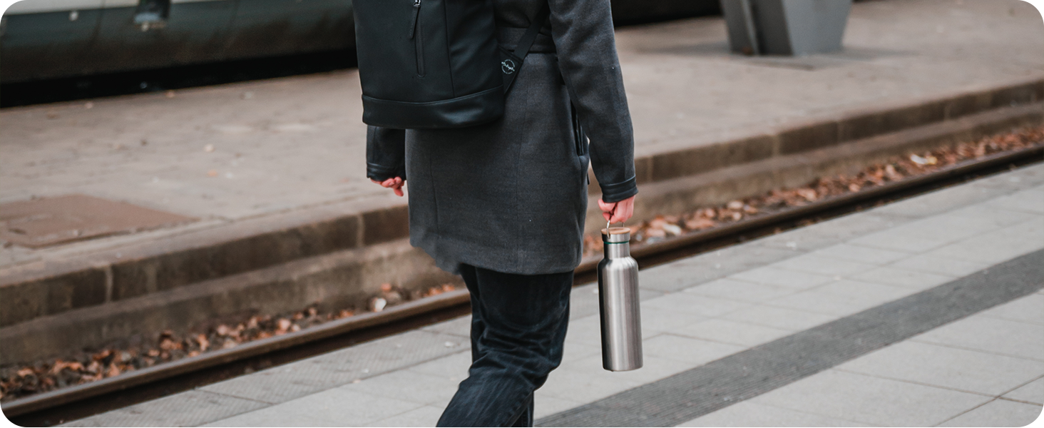 Person in dark coat walking on a platform or sidewalk next to train tracks. Lower body and legs visible, carrying a bag or briefcase.