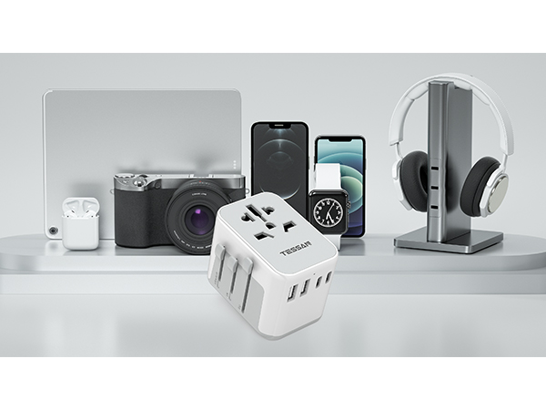 Display of electronic devices: camera, smartphones, wireless earbuds, multi-port USB charger, and over-ear headphones with charging stand on a gray surface.