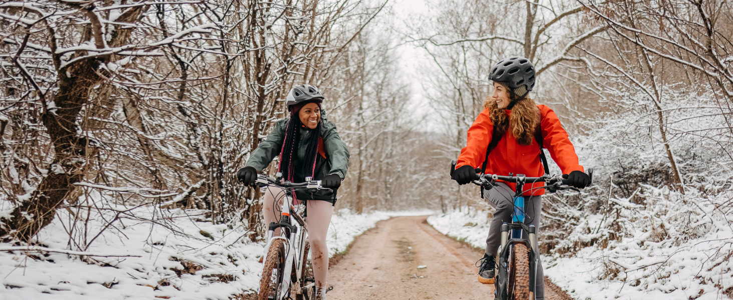 Two cyclists riding bicycles on a snowy path through a winter forest. One wears a red jacket, the other a dark jacket.