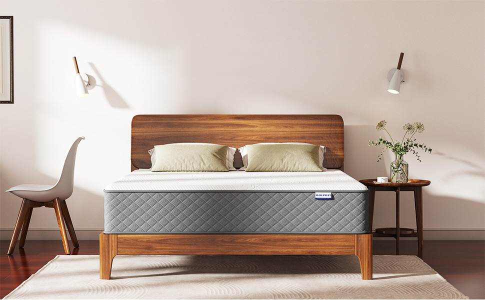 Queen-size bed with wooden frame and headboard, gray quilted mattress, white pillows. Flanked by nightstands with lamps, modern chair in foreground.