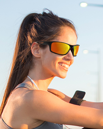 Woman wearing orange-tinted sports sunglasses outdoors. A small device is visible on her upper arm.