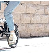 Vista de cerca de piernas y pies sobre pedales de bicicleta, que muestra jeans y calzado negro sobre un fondo de pared de piedra clara