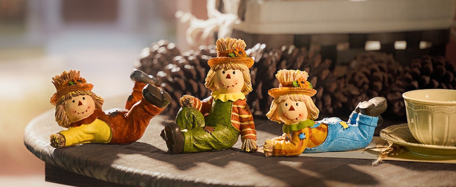 Three small decorative figurines of smiling characters in colorful outfits, lying on their stomachs. Figures have troll-like features with wild hair and are positioned on a dark surface near a white tea cup.