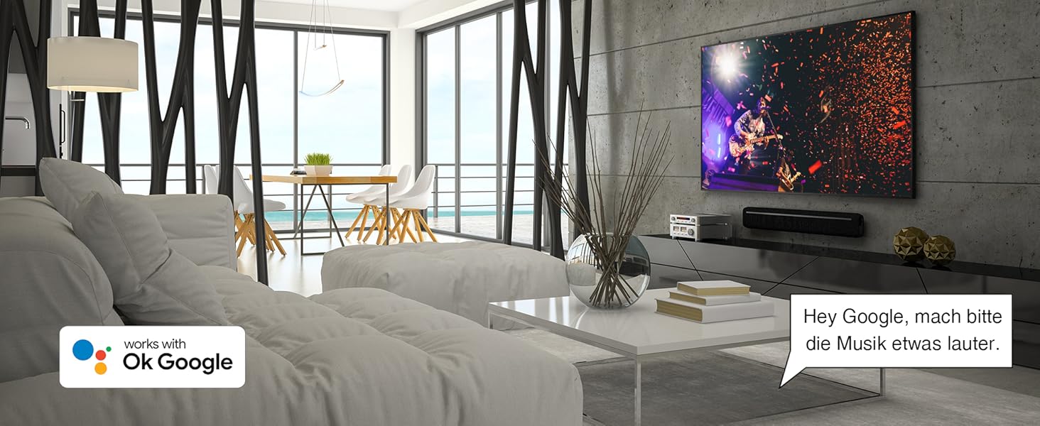Modernes Wohnzimmer mit großen Fenstern, grauem Sofa und Wandfernseher. Google Assistant-Logo in der Ecke