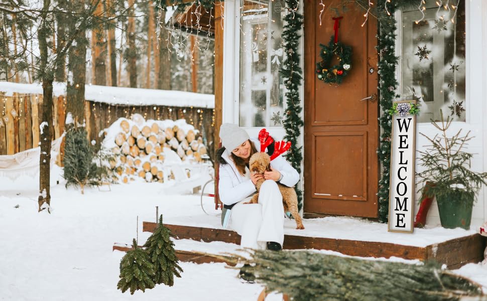 Woman in white with dog in antlers by snow-covered door with &#34;WELCOME&#34; sign and wreath.