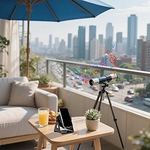 Balcony setup with telescope, blue umbrella, and wooden table holding drinks and phone stand. City skyline with high-rise buildings visible in background.
