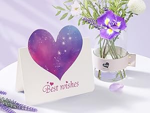 Weiße Grußkarte mit violettem Galaxie-Herzdesign neben einer Glasvase mit lila Blumen und grünem Laub.