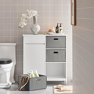 White bathroom storage cabinet with grey fabric drawers. Features orchid vase, toiletries on top. Positioned against tiled wall near toilet.