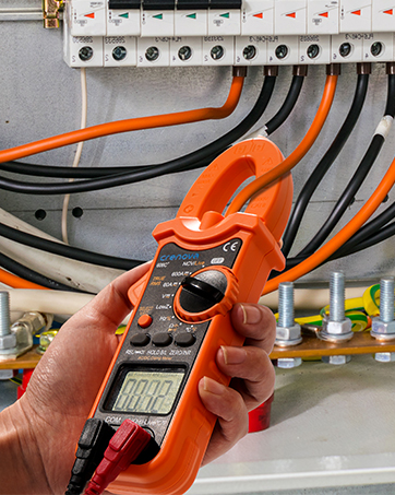 Hand holding an orange clamp meter near electrical wiring and circuit breakers. The device is being used to measure electrical current.