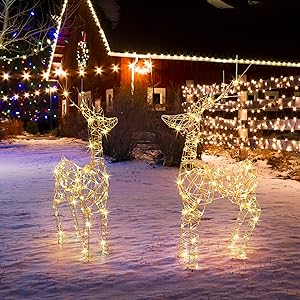 Two illuminated wire frame reindeer decorations on snow-covered ground. Festive lights adorn a building in the background, creating a holiday atmosphere.