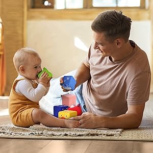 A young father smiled and looked at his baby, who was sitting on the carpet chewing on soft blocks