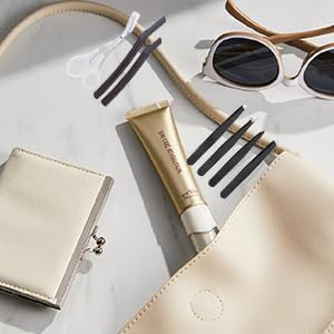 Flat lay of beauty accessories: sunglasses, makeup brushes, golden tube (likely cosmetic product), white plate, and light-colored pouches on neutral background.