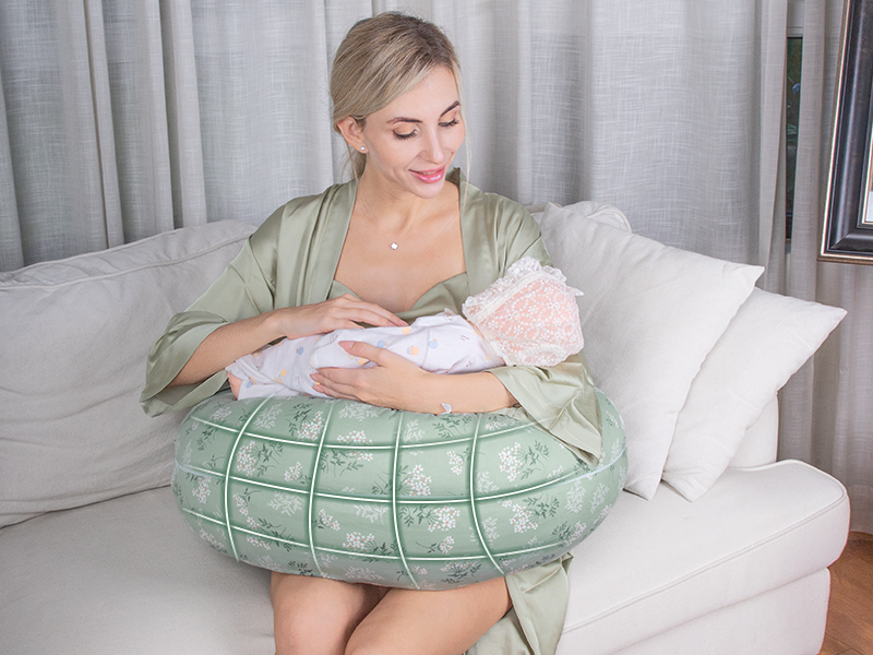 Frau in grüner Robe sitzt auf einem gemusterten Kissen, hält ein Kind und schaut es an. Szene in einem hellen