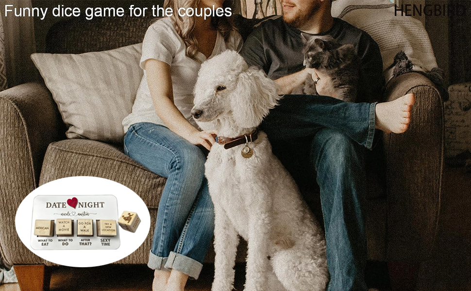 Couple on couch with two large dogs. Dice game box visible in foreground. Casual indoor setting with throw pillows and blanket.