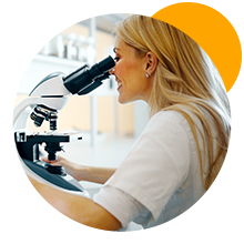 Une femme aux longs cheveux blonds vêtue d'un haut blanc, regardant au microscope dans un laboratoire. Une grande forme circulaire jaune est visible à l'arrière-plan.