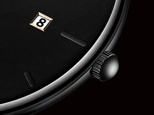 Close-up of a minimalist black watch face with silver hands and a small date window showing '8'. A curved edge and crown are visible against a dark background.