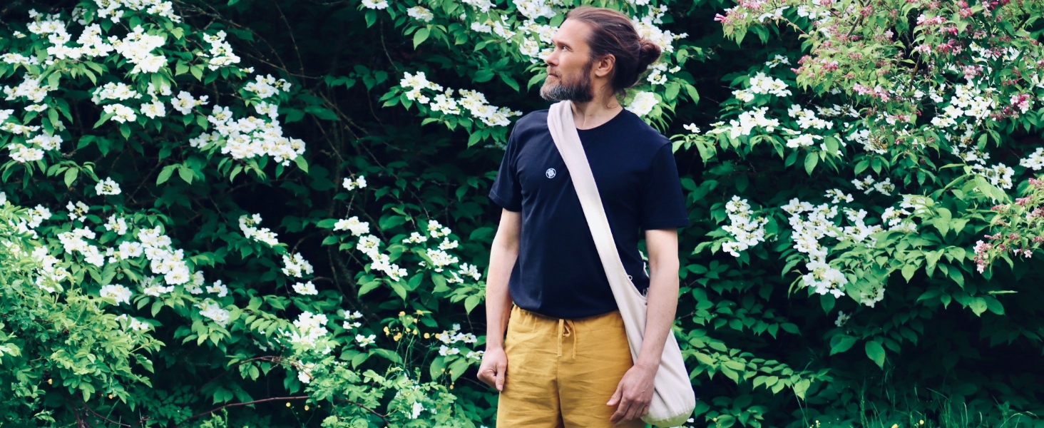 Mann in dunklem Hemd und gelber Hose, der vor blühenden Büschen steht und eine helle Schultertasche trägt